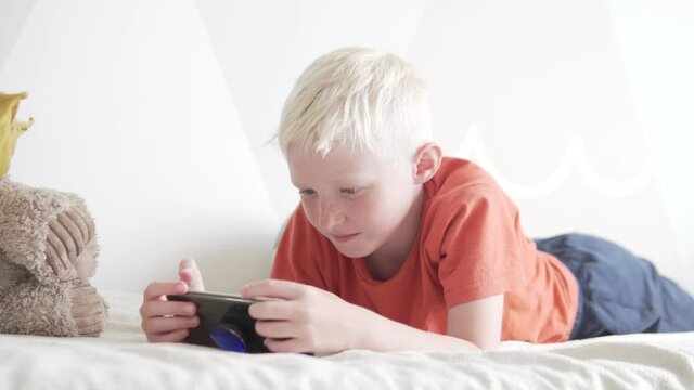Beautiful blonde boy plays the game on his mobile phone at home on the bed.
