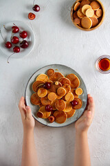 Tiny pancakes with cherries on a plate, children's hands, top view. - obrazy, fototapety, plakaty