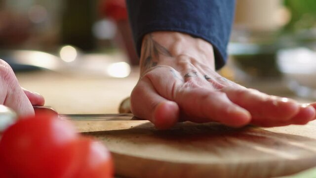Close-up shot of hands of male cook crushing garlic clove with knife and then trimming the root end and peeling it above wooden cutting board