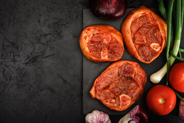 raw pork shank barbecue steaks in a marinade with fresh vegetables, spices, onions and garlic on a stone cutting board on a black concrete background. top view. artistic layout with copy space