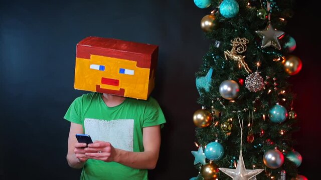 A man in a square mask uses a smartphone. He presses his hands on the display of the phone. Nearby is a Christmas tree.
