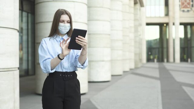 Young Businesswoman With Medical Mask Is Working On Her Tablet Then Look Up And Return To Work Again On The Street Near Modern Office Building During Pandemic Covid-19 Coronavirus Quarantine.