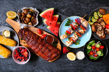 BBQ or summer picnic food table scene. Selection of grilled meats, vegetables, fruits, salad and potatoes. Above view over a dark stone background.