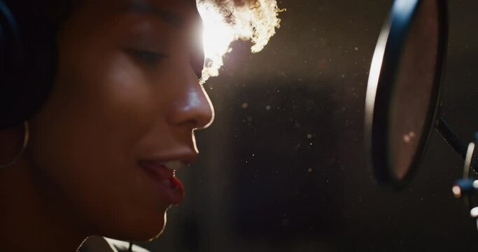 Close up of an young professional smiling energetic african female singer wearing headphones is performing a new song with a microphone while recording it in a music studio. 
