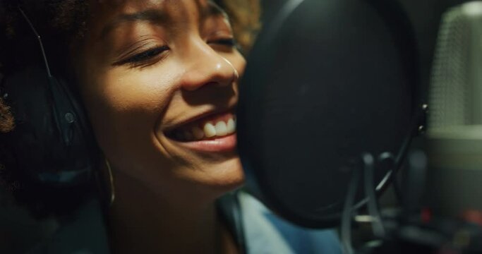 Close up of an young professional smiling energetic african female singer wearing headphones is performing a new song with a microphone while recording it in a music studio. 