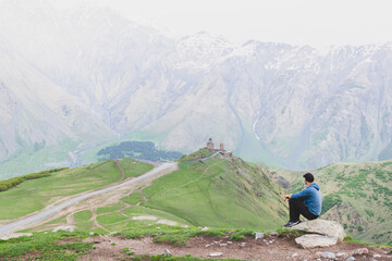 Fototapeta na wymiar Male tourist in casual clothes with sits relaxed and looks to Gergeti trinity church surounded by green nature. Solo travel in Kazbegi national park.