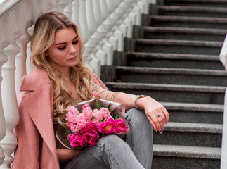 Woman in pink topcoat sits on steps with pink flowers bouquet in arms .