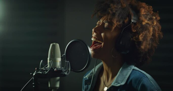 An young professional smiling energetic african female singer wearing headphones is performing a new song with a microphone while recording it in a music studio. 