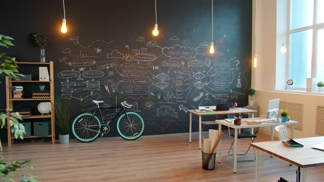 Zoom-out of modern creative office with blackboard wall and desks with no people illuminated with lights lamps. Interior, workplace and business concept.