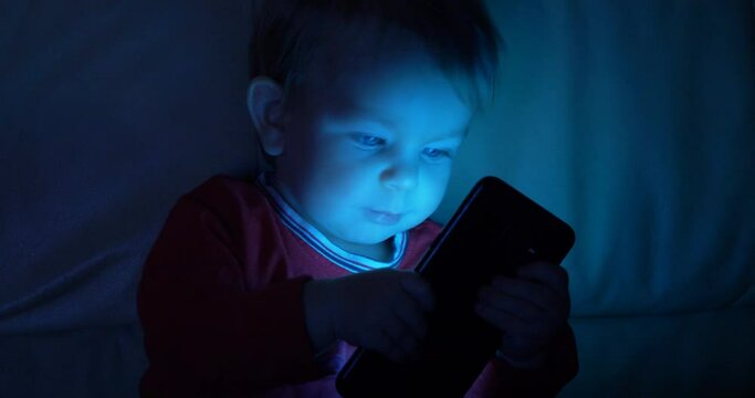 Portrait of baby child look at mobile phone tin dark night, too early technology addiction