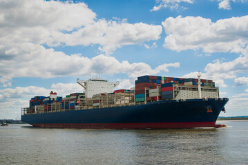 A cargo ship with many colorful containers floats along the river on a sunny day.