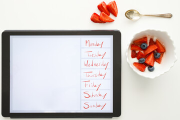 Bowl with strawberries, blueberries and yogurt and electronic tablet with the days of the week on a white background. Concept of healthy breakfast and productivity.