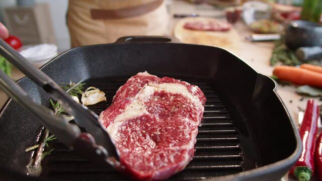 Zoom in shot of male chef using metal tongs while placing raw meat steak on hot grill pan with rosemary and garlic cloves on it