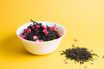 a bunch of tea with dried strawberries in a small white bowl on a yellow background, a handful of tea is scattered on a yellow background