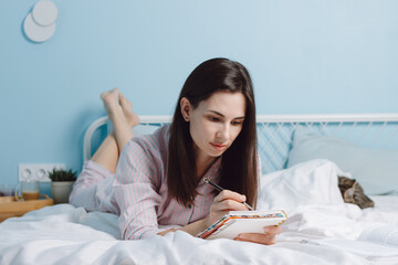 Obraz na płótnie Canvas Portrait of brunette attractive lovely charming creative cheerful girl lying in bed with cat, creating story, making notes in notepad in modern white light bedroom interior. Good wake up concept