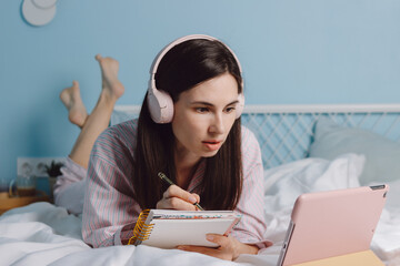 Close up side view of young motivated calm brunette millennial woman in headphones lying on bed and watch webinar using digital tablet. Girl with bated breath listen to tutor, coach, making notes