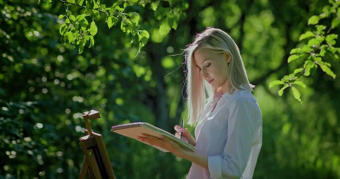 Drawing on the nature. Girl draws in an album, makes a sketch. Portrait of a beautiful girl painting not on nature in an album.