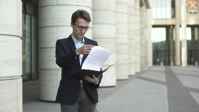 Young businessman in dark blue suit and glasses check documents near modern office building and laught and smile while cheering.