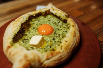 Khachapuri Made From a Delicious Tender Dough with Spinach, Melted Cheese and Butter Close Up. Traditional Hachapuri Topped with Spinacia Leaves on Wooden Rustic Background