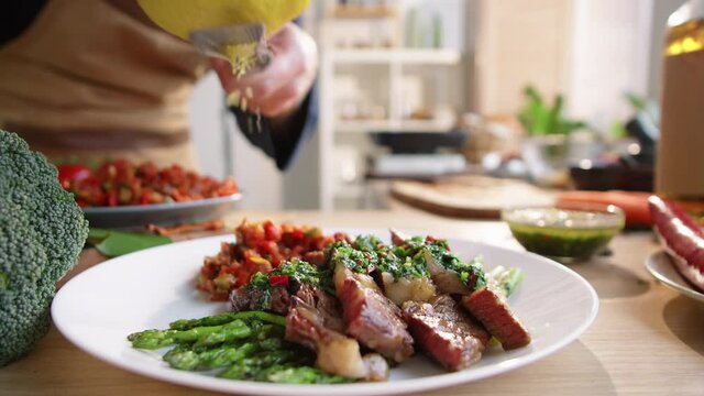 Close up shot of professional male chef grating lemon peel on roasted meat served on white plate with asparagus, vegetables and green sauce