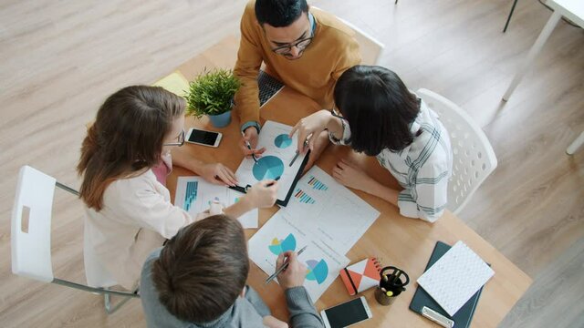 Group of office workers Caucasian and Middle Eastern are talking and pointing at charts on paper sitting at desk in office. Discussion and employees concept.