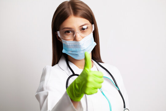 Young female doctor in a mask looks at the camera and holds a thumb up