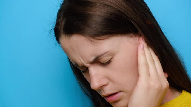 Close up of stressed frowning young woman plugging ear, feeling painful discomfort, avoids bad sound, models against blue studio background. Unhealthy brunette girl suffering from strong earache