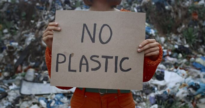 Crop view of woman raising carton poster with no plastic writing. Female person protesting against earth pollution while standing in front of trash hill. Concept of ecology