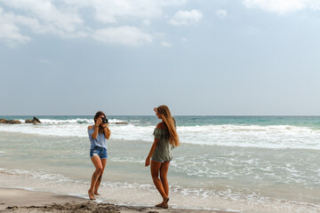 Fototapeta premium Traveling and photography. Young woman with camera a taking picture of her friend on the sea beach