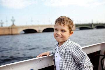 Fototapeta premium cute caucasian boy looking aside wearing henley tee and shirt on a river boat tour along rivers and channels of Saint Petersburg. Trinity Bridge on background. Image with selective focus 