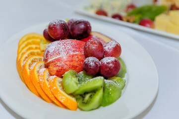 Fruit assortment of apples, oranges, kiwis and grapes on a white plate, close-up. Dessert snacks on banquet table.