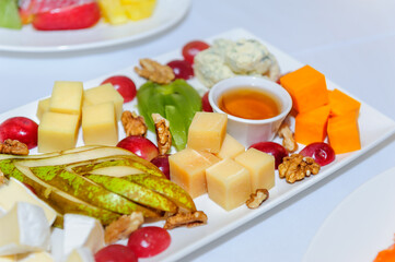 Cheese plate at banquet, close up. Assorted different types of cheeses with honey decorated with nuts, pear, kiwi, grape on large white dish.