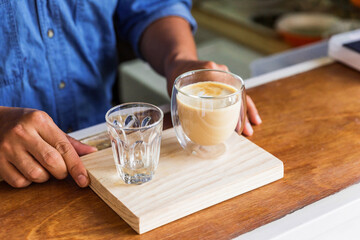 Male barista serve hot latte coffee and fresh water in drinking glass at the wooden counter bar. Served on wooden plate.