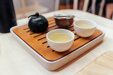 Close-up of green tea set in cups on small wooden plate with kettle and strainer.