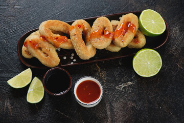 Fried panko breaded calamari rings with lime and dips over dark brown stone background, studio shot