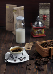 A cup of hot black coffee, coffee beans in a wooden box and chocolate on a dark wooden table. Bottle of milk and manual coffee grinder.