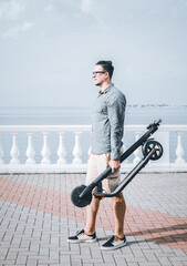 Young man standing with folding electric scooter on city embankment in front of sea.