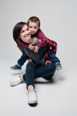 Smiling beautiful brunette mom with little son, sitting on floor at studio, embracing and looking at camera. Parent holding on shoulder her child in checked shirt and jeans. Happy relationship.Gray