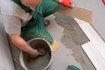 The builder applies cement adhesive to the ceramic tile with a stainless trowel.