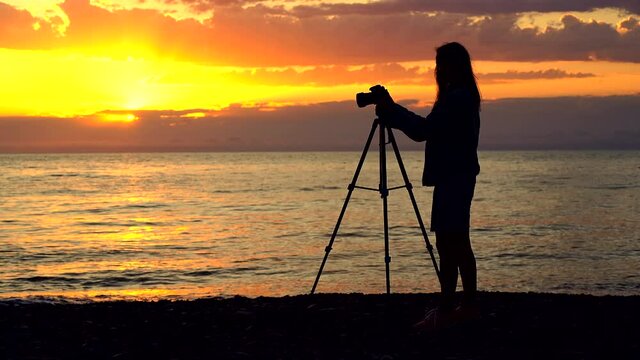 Silhouette of a woman photographer taking a photo of the sea with professional dslr camera on tripod at sunset 