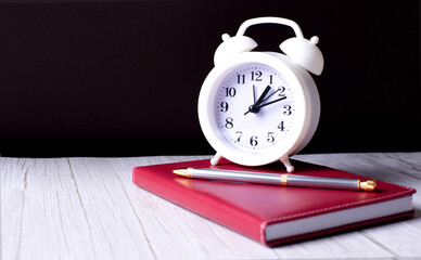 On a light wooden table is a burgundy notebook with a pen and an alarm clock