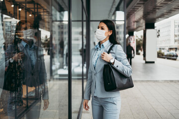 Fototapeta premium Young and elegant business woman with face protective mask standing on empty street and looking at storefront of closed store. Corona virus or Covid-19 concept.