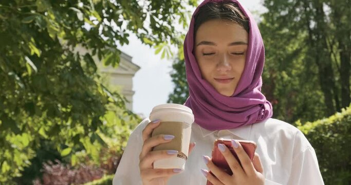 Beautiful young woman drink coffee and looking at smartphone, wearing traditional headscarf. Attractive Female in hijab