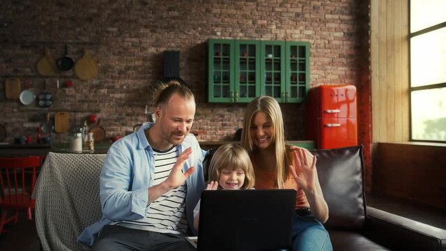 Parents and son in casual clothes smiling, enjoying online video call on laptop computer, sitting on couch in studio apartment with stylish interior