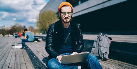Portrait of caucasian male graphic designer editing project on laptop while sitting outdoors during sunny autumn day, handsome hipster guy dressed in trendy apparel spending time with netbook