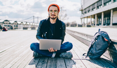 Portrait of positive caucasian male student in eyewear sitting on wood and looking at camera while...