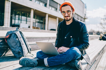 Portrait of smiling trendy dressed male student in spectacles using laptop computer for networking...
