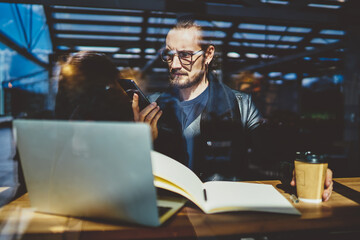 Puzzled male freelancer in optical spectacles for vision protection reading received email message during internet browsing, confused hipster guy updating smartphone during distance job in coffee shop