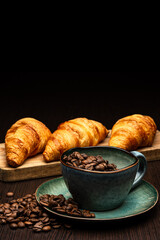 Cup with coffee beans on a black background. Freshly roasted arabica. Several fresh croissants on a wooden board.
