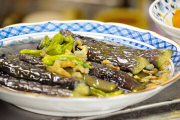 おばんざい屋さんのカウンターに並んだ、白い陶器の丼鉢に盛り付けられた、なすの揚げ浸しと添えられた海老とししとう がらし。
Some deep fried eggplant & shrimp & green pepper in Japanese broth in the earthenware donburi boul on the counter table.